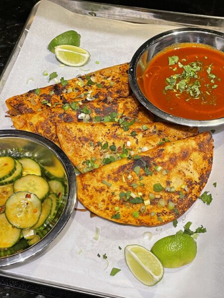 Four tacos on a baking tray lined with paper, alongside a bowl of sliced cucumbers and consumé, with limes arranged on the side