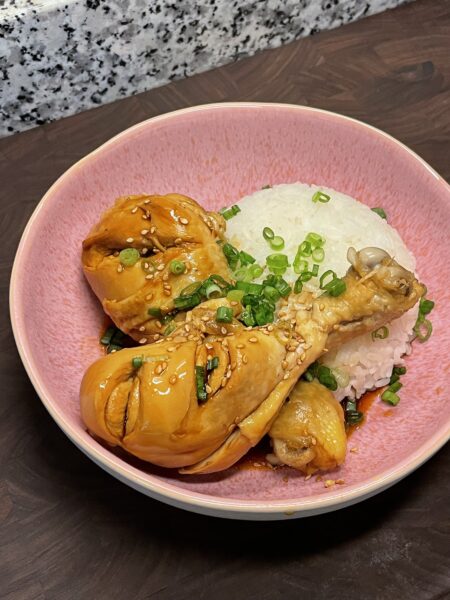 Two chicken drumsticks, covered in a thickened soy sauce, arranged next to a pile of rice in a pink bowl