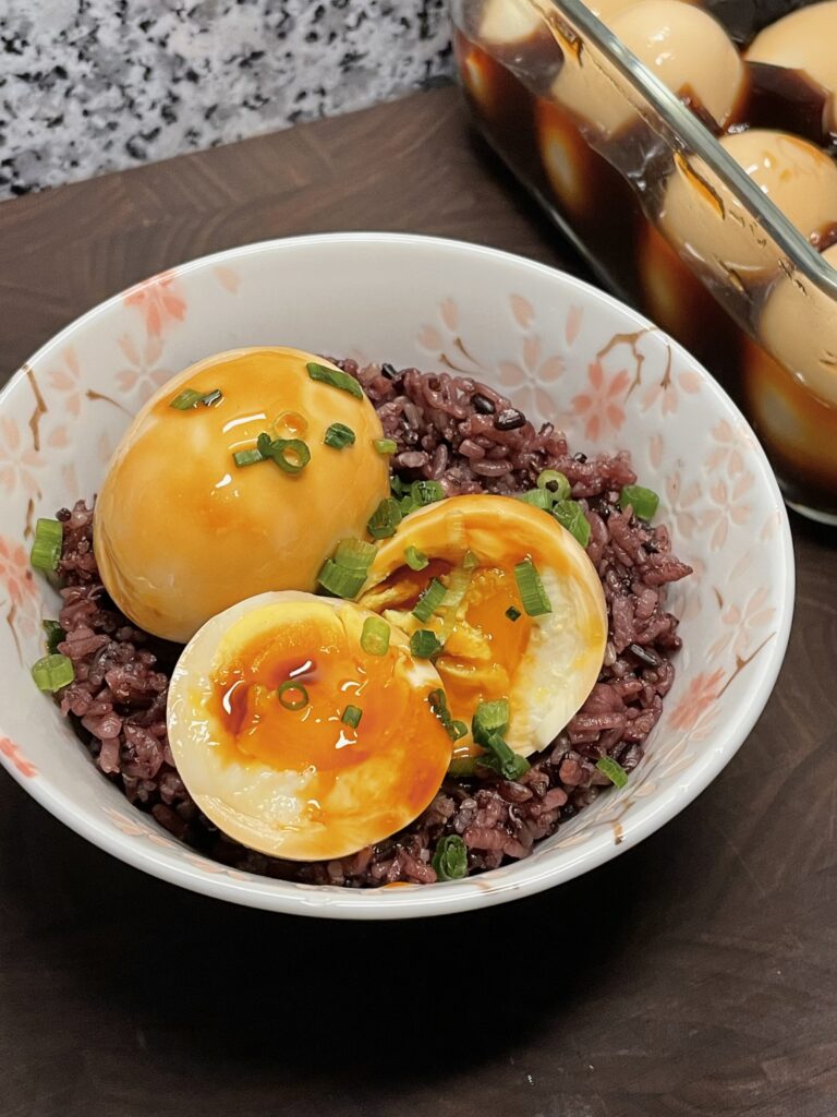 Two marinated eggs, one with a jammy yolk, sitting on a bed of multigrain ("purple") rice, garnished with scallions