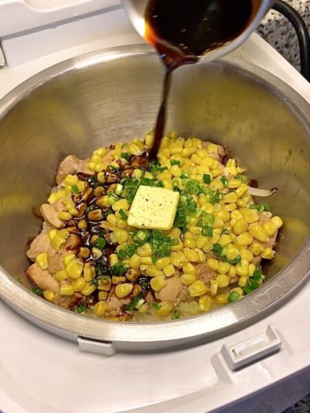 Rice cooker bowl with chicken, corn, scallions, a knob of butter, and black pepper, with a brown sauce being poured in