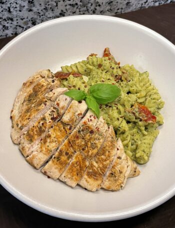 Wide bowl of pesto pasta (fusilli) with sun-dried tomatoes, with sliced chicken breast on top, garnished with chili flakes and basil