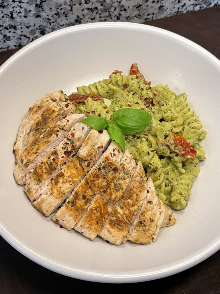 Wide bowl of pesto pasta (fusilli) with sun-dried tomatoes, with sliced chicken breast on top, garnished with chili flakes and basil