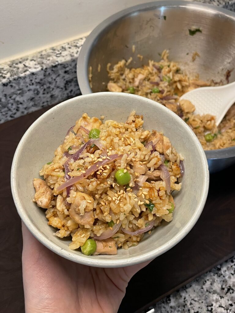 Hand holding a bowl of rice with pieces of chicken, sliced red onion, and green peas, garnished with sesame seeds