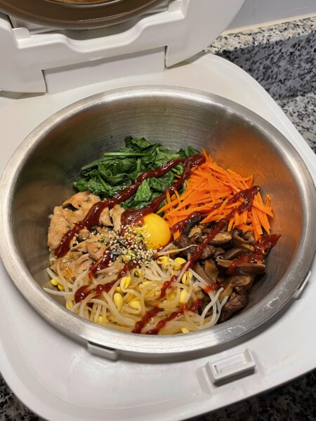 Rice cooker bowl with chicken, spinach, carrots, mushrooms, bean sprouts, and an egg yolk, topped with spicy red sauce and seaweed flakes