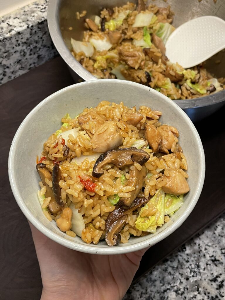 Hand holding a bowl of rice mixed with chicken pieces, sliced mushrooms, napa cabbage, goji berries, and scallions
