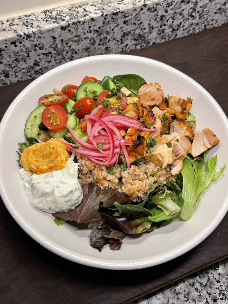 Shallow bowl with salad greens, topped with grain, chopped chicken thigh, cucumber and tomato mix, red pepper hummus, tzatziki (yogurt sauce), pickled red onions, and freshly chopped parsley