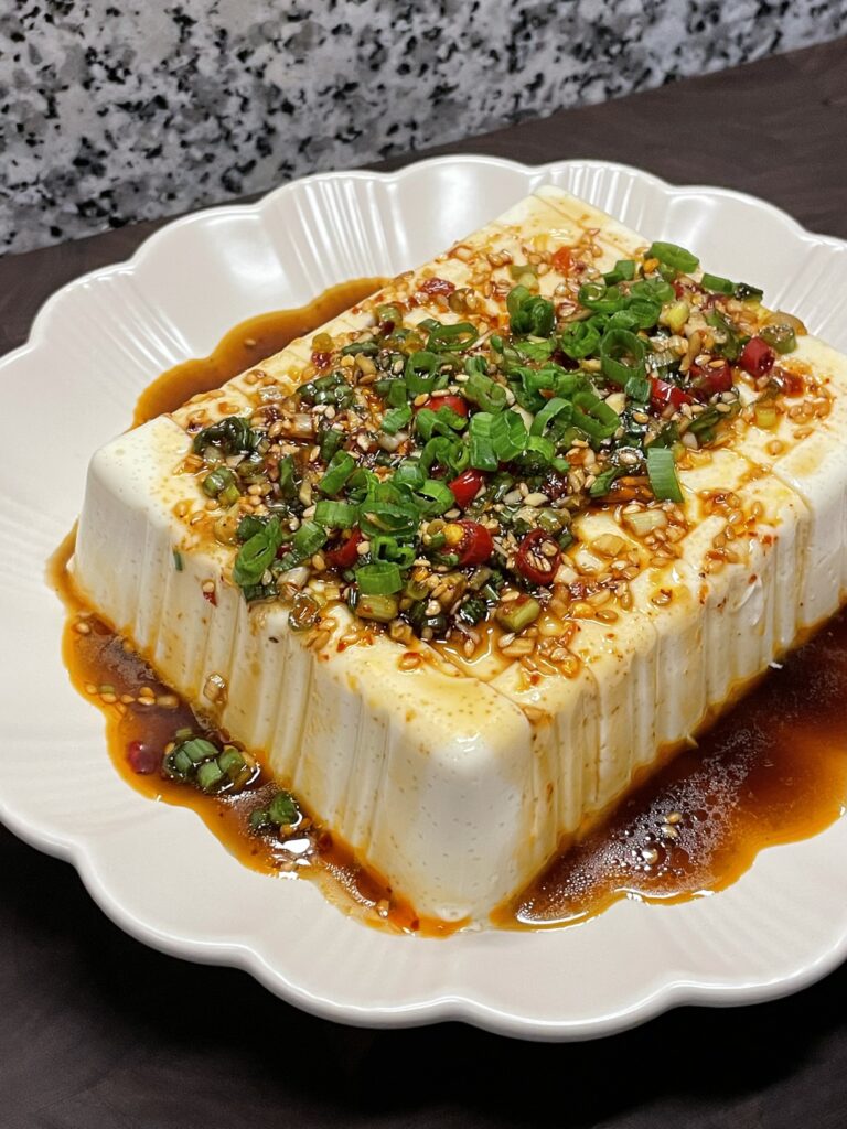 Block of tofu on a circular plate, drizzled with a spicy soy sauce, sesame seeds, and chopped scallions (green onions)