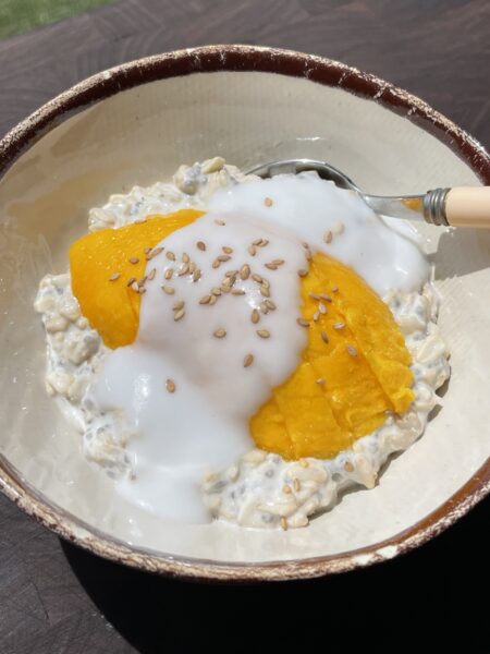 Bowl of overnight oats under direct sunlight, topped with sliced fresh mango, thick white coconut sauce, and toasted white sesame seeds