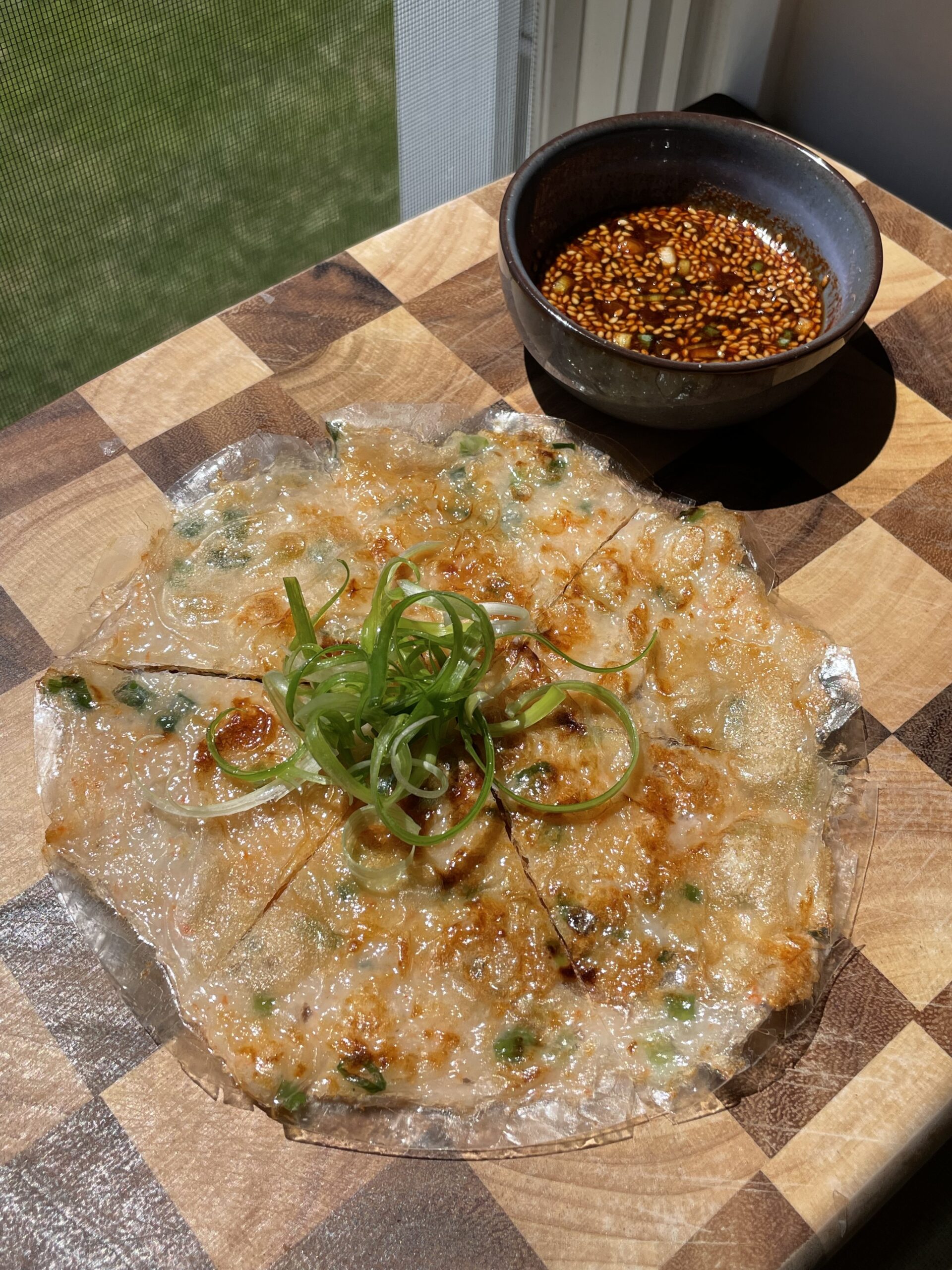Golden-brown, crispy pan-fried pancake on a checkered cutting board, garnished with curled scallions (green onions), with a small blue bowl of spicy soy sauce on the side