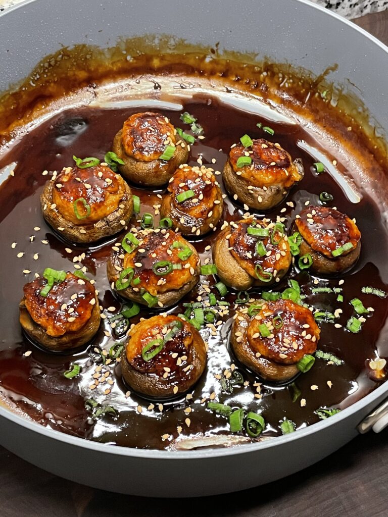A pan with mushrooms filled with shrimp paste, on a shiny brown sauce and garnished with chopped scallions (green onions)