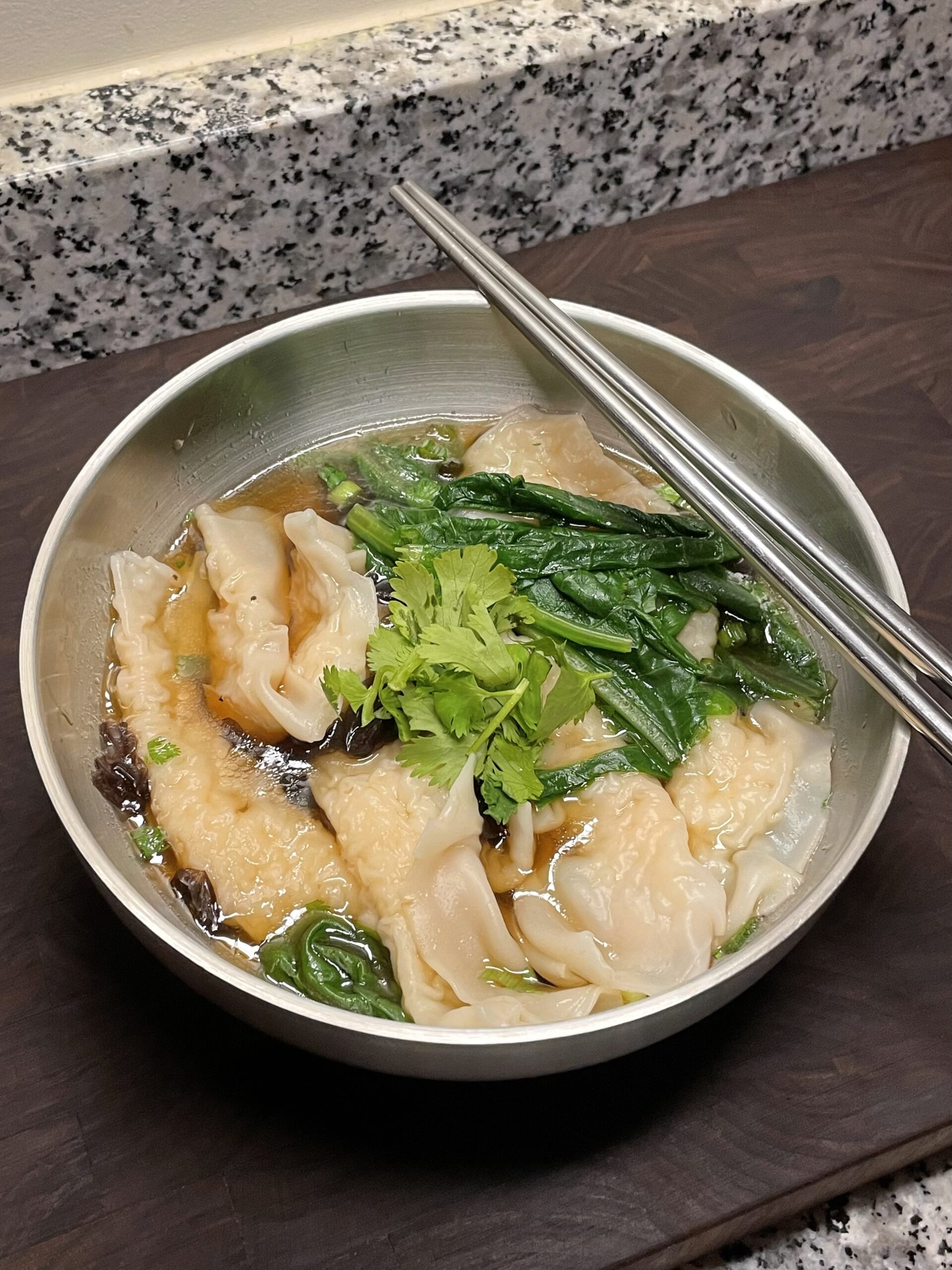 Stainless steel bowl of noodles filled with shrimp paste, blanched leafy greens, and garnished with cilantro, in a soupy broth
