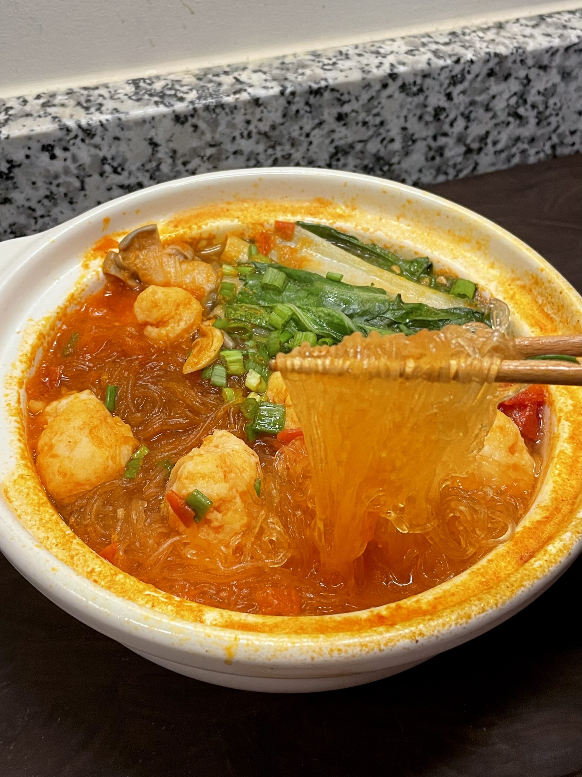 White clay pot with tomato broth, shrimp balls, vermicelli, and blanched leafy greens, garnished with chopped scallions (green onions)