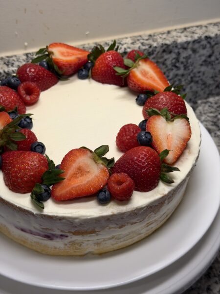 Overhead close-up of a naked berry chantilly cake with whipped cream, topped with a ring of fresh strawberries (halved), raspberries, and blueberries, set on a white cake stand.