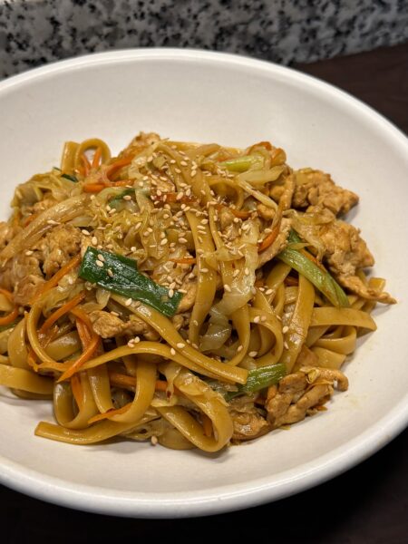 A white bowl of chicken chow mein with wide flat noodles, shredded chicken, cabbage, carrots, and scallions, garnished with sesame seeds.