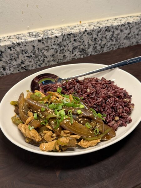 A white oval plate with stir-fried chicken and glazed eggplant strips topped with scallions and sesame seeds, served alongside purple rice.