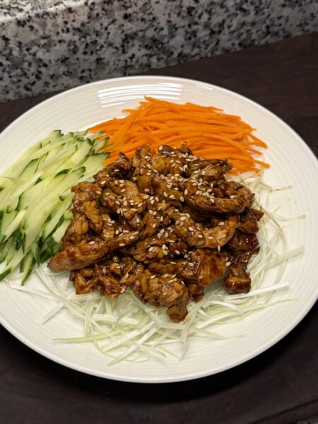 A white plate of glazed, sesame-topped shredded chicken served with piles of julienned cucumber, carrot, and scallion.