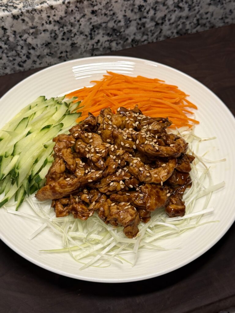 A white plate of glazed, sesame-topped shredded chicken served with piles of julienned cucumber, carrot, and scallion.