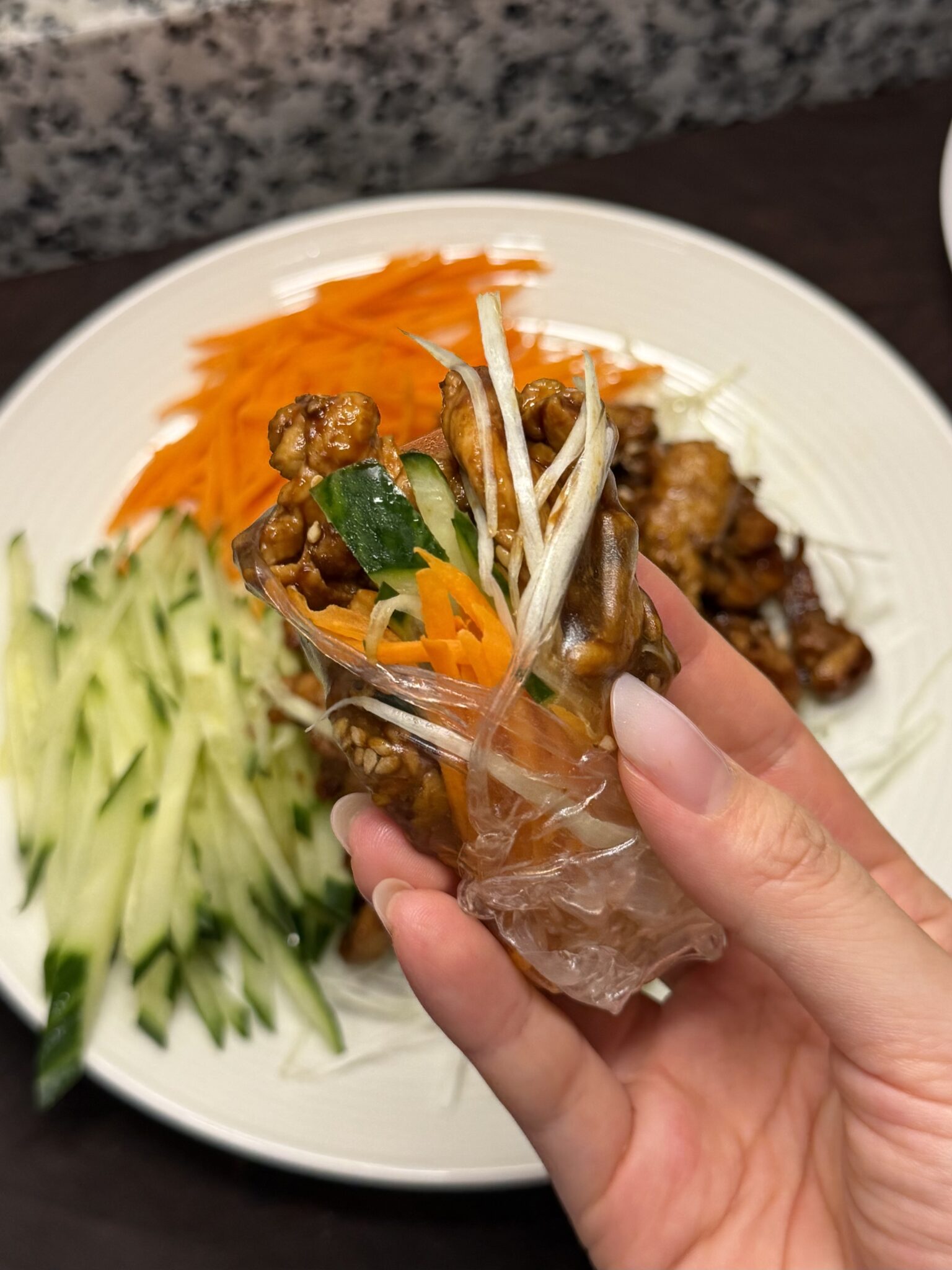 A hand holding a thin rice wrapper filled with glazed shredded chicken, cucumber, carrot, and scallion, rolled up over a plate of the same ingredients.