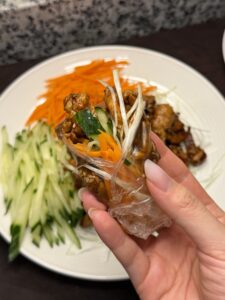 A hand holding a thin rice wrapper filled with glazed shredded chicken, cucumber, carrot, and scallion, rolled up over a plate of the same ingredients.