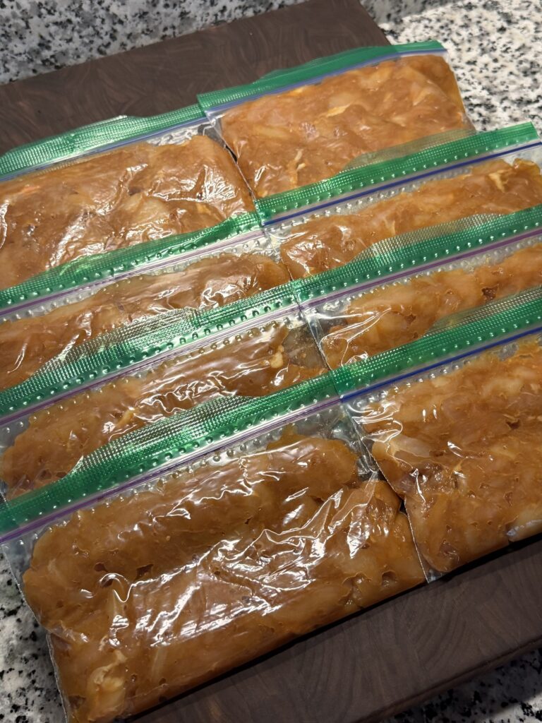 Several ziplock bags filled with marinated, shredded chicken breast, flattened and stacked in rows on a wooden cutting board.