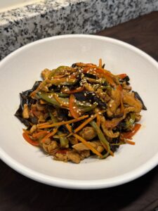 A white bowl of fish-fragrant shredded chicken stir-fried with wood ear mushroom, carrots, and green and red peppers, topped with sesame seeds.