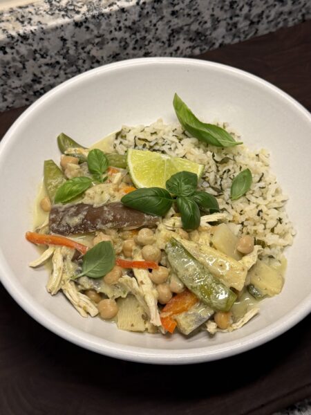Shallow white bowl of chickpea Thai green curry with vegetables, herbed rice, basil, and a lime wedge.