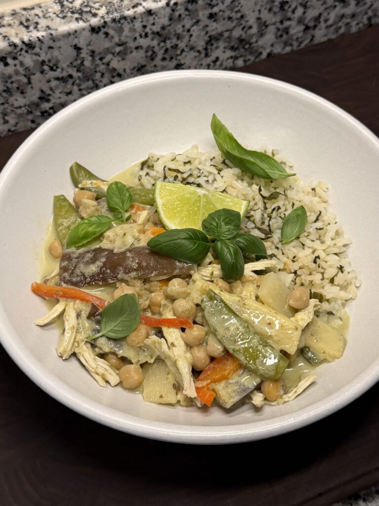 Shallow white bowl of chickpea Thai green curry with vegetables, herbed rice, basil, and a lime wedge.