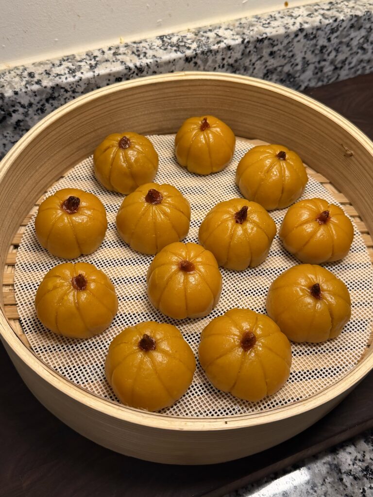 Twelve steamed pumpkin mochi in a bamboo steamer basket, each shaped like a tiny pumpkin with vertical ridges and goji berry "stems" on top.