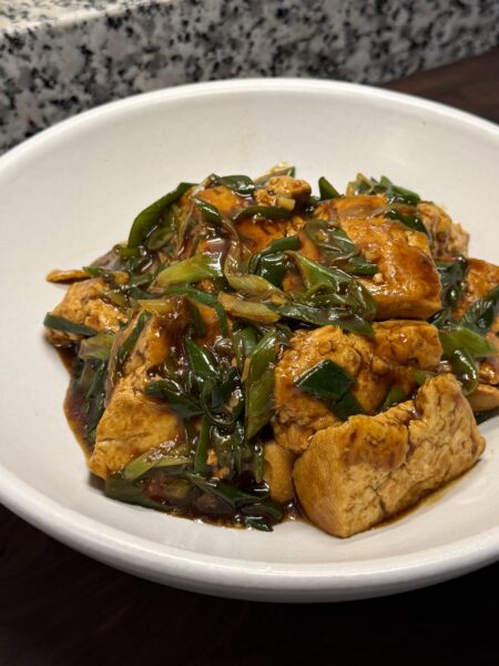 Soy braised tofu in glossy brown sauce, topped with lots of sliced green scallions, in a shallow white bowl.