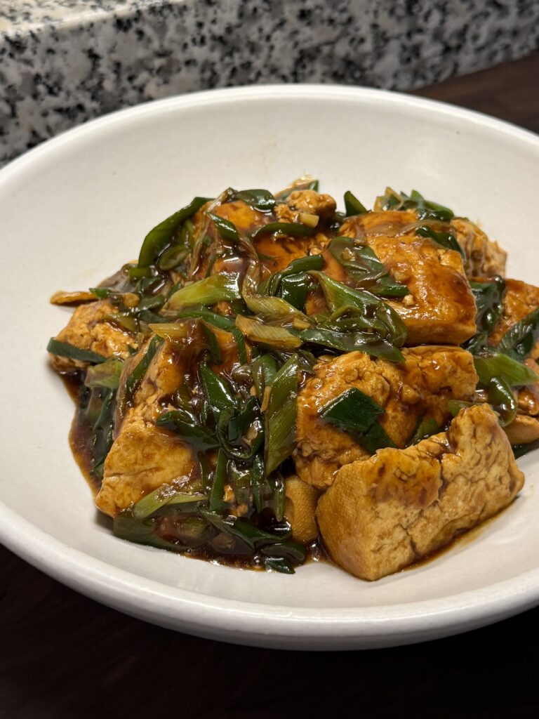 Soy braised tofu in glossy brown sauce, topped with lots of sliced green scallions, in a shallow white bowl.